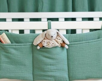 May include: A sage green fabric crib organizer with three pockets hanging from a white wooden crib. The left pocket holds a wooden brush, and the center pocket contains a small plush rabbit toy with a lace collar. The organizer is made of a textured muslin material.