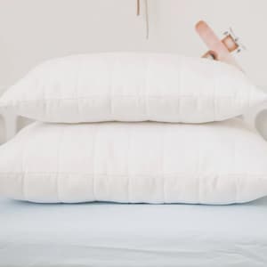 May include: Two rectangular white pillows stacked on top of each other, featuring a quilted grid pattern. The pillows are positioned on a light blue sheet within a white frame, with a beige balloon-shaped wall hanging and a bear-shaped rainbow decoration.