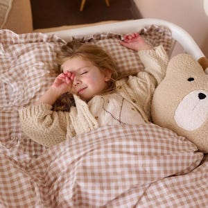 Op de afbeelding: Een beige en wit beddengoedset met een vichy-ruitpatroon, inclusief een kussensloop met ruches en een bijpassend dekbedovertrek. Een beige pluche kussen in de vorm van een beer met een witte snuit en zwarte geborduurde ogen en neus ligt op het bed.