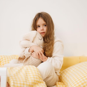 May include: A plush cream-colored stuffed rabbit toy with embroidered closed eyes and long ears, held by a person wearing a cream-colored long-sleeved top. The toy features a soft, textured fabric body and a smooth, knit-like fabric on its lower section.
