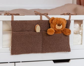 May include: A dark brown, textured sherpa fabric storage organizer with two pockets, attached to a white crib rail with brown ribbon ties. One pocket contains a small wooden hairbrush, and the other holds a plush brown teddy bear.