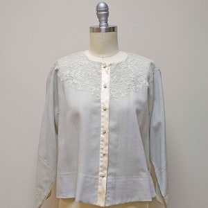 Op de afbeelding: Een lichtblauw blouse met een witte kanten kraag en lange mouwen. De blouse heeft een knoopsluiting aan de voorkant en een satijnen bies.