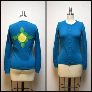 Puede incluir: Un cárdigan azul turquesa con un diseño de flor verde y amarilla en la espalda. El cárdigan está en un maniquí blanco.