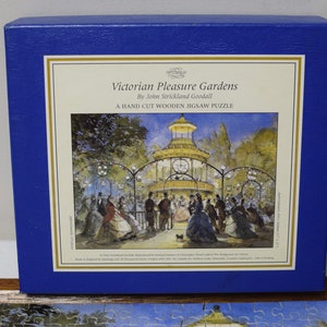 May include: A blue box with a hand-cut wooden jigsaw puzzle of a Victorian Pleasure Garden by John Strickland Goodall. The image shows a bandstand with people dancing and a dog.