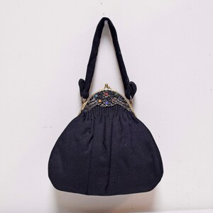 Puede incluir: Un bolso de tela negro con cierre dorado y joyas de colores. El bolso tiene un solo asa.