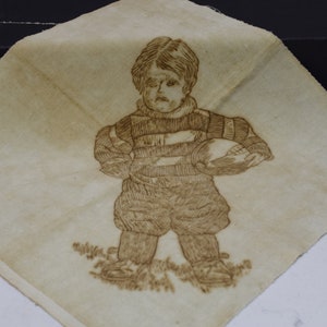 May include: A vintage embroidered fabric square featuring a child in a striped shirt holding a football. The fabric is a light beige color with brown embroidery.