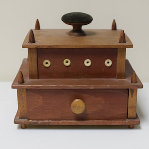 May include: A wooden sewing box with a drawer and a pincushion on top. The box has a brown stain and is decorated with small wooden pegs. The drawer has a wooden knob.
