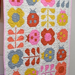 Puede incluir: Un edredón hecho a mano con un patrón repetido de mariquitas, flores y formas de hojas de colores sobre un fondo blanco. El edredón tiene un borde rosa y está colgado de una percha de madera.