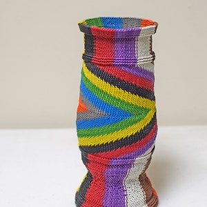 May include: A colorful woven vase with a striped pattern in shades of red, purple, white, black, yellow, green, and blue. The vase has a wide base and a narrow neck.