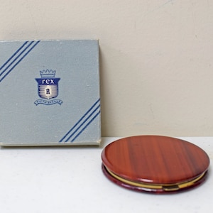 May include: A vintage compact mirror with a wood-grain design on the lid. The lid is closed and the mirror is sitting on a white surface. The compact mirror is in a blue box with the text "Rex" and a crown.