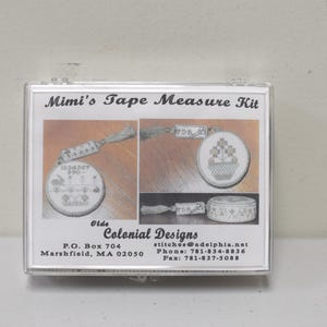 Może przedstawiać: Przezroczyste plastikowe etui zawierające zestaw miary taśmy. Zestaw zawiera miarę taśmy z kwiatowym wzorem i tekstem "Mimi's Tape Measure Kit". Zestaw pochodzi od Olde Colonial Designs, z siedzibą w Marshfield, MA.