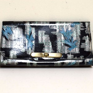 Könnte beinhalten: Eine schwarze und silberne Clutch mit einem goldenen Verschluss. Die Clutch hat ein blaues und weißes abstraktes Muster.