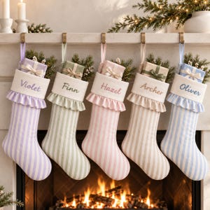 Op de afbeelding: Vijf hangende kerstsokken met pastelkleurige strepen in lavendel, saliegroen, roze, beige en lichtblauw. Elke sok heeft een witte boord met een geplooide rand, gepersonaliseerd met de geborduurde namen Violet, Finn, Hazel, Archer en Oliver.