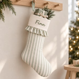 Könnte beinhalten: Ein Weihnachtsstrumpf mit hellgrün-weißem Längsstreifenmuster und einem cremefarbenen Umschlag. Der Umschlag ist mit dem Namen Finn in grünem Garn bestickt und mit einer Rüschenkante versehen. Ein kleines Geschenk steckt im Strumpf.