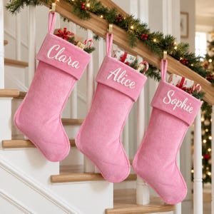 Op de afbeelding: Drie zachte roze kerstsokken hangen aan een houten trapleuning. Elke sok is voorzien van een wit geborduurde naam op de boord: Clara, Alice en Sophie. De sokken zijn gevuld met kleine ingepakte cadeautjes en zuurstokken.