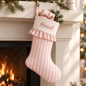 May include: A Christmas stocking featuring a white cuff embroidered with the name Hazel in pink script. The stocking has a pink and white striped pattern, a ruffled trim below the cuff, and contains a small gift box with a pink ribbon.