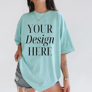 May include: A pale blue t-shirt with the words "YOUR Design HERE" printed in black. The shirt has a relaxed fit. The model is wearing denim shorts and a gold necklace. The t-shirt is made of a soft material.