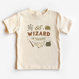 May include: Cream-colored t-shirt with a brown and cream graphic that reads "WIZARD IN TRAINING." The design includes illustrations of potion bottles, glasses, a frog, a wand, an owl, and a cauldron. The shirt is made of soft material.