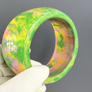 May include: A wide bangle bracelet featuring a marbled pattern of vibrant green, bright yellow, and soft pink colors. The smooth, polished surface of the circular accessory displays a swirling, multi-colored design that is consistent throughout the piece.