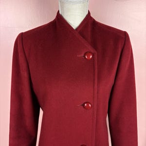May include: A deep red coat featuring a high collar and an asymmetrical front closure. The garment is secured with two visible, round, matching red buttons positioned along the vertical edge of the overlapping front panel.