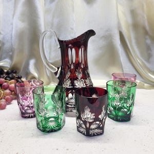 Puede incluir: Una jarra de cristal tallado rojo rubí con un asa transparente y seis vasos a juego en tonos rojos, verdes y rosas. La cristalería presenta un diseño floral detallado. El conjunto está dispuesto sobre una superficie blanca.