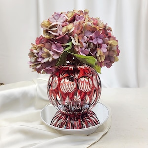 May include: A round, deep red cut glass vase featuring intricate geometric patterns and clear glass accents. The vase is filled with a large, dense bouquet of artificial hydrangea flowers in shades of dusty purple, mauve, and muted green.