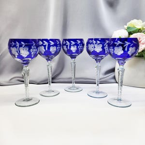 Op de afbeelding: Vijf kobaltblauwe wijnglazen met geëtste bloemen- en druivenontwerpen. De glazen hebben heldere, getextureerde stelen en ronde bases. De achtergrond is een zachte grijze stof en een vaas met bloemen.
