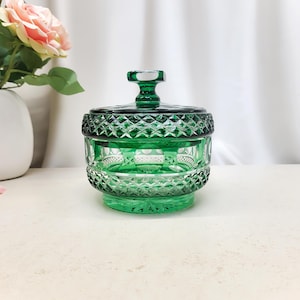 Op de afbeelding: Een ronde smaragdgroene glazen snoeppot met bijpassend deksel. De buitenkant is voorzien van een gedetailleerd geslepen patroon met ruitvormige facetten rond de rand en basis, en een gladde, cirkelvormige band in het midden van de kom.