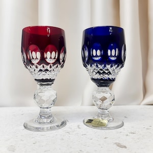 May include: Two crystal wine glasses, one with a deep red bowl and one with a deep blue bowl. Both feature a clear, faceted stem and base, with a diamond-cut pattern on the lower bowl and circular indentations around the upper rim of the glass.