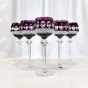 Op de afbeelding: Een set van vijf kristallen wijnglazen met dieppaarse kelken, voorzien van een geslepen ruitpatroon aan de onderzijde en cirkelvormige inkepingen bij de rand. Elk glas heeft een slanke, gefacetteerde steel en een brede, ronde voet.