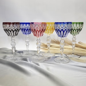 May include: Six colorful crystal wine glasses. Each glass features a different colored bowl: black, blue, pink, yellow, blue, and green. The stems are clear with a decorative ball design. The glasses are arranged on a white surface.