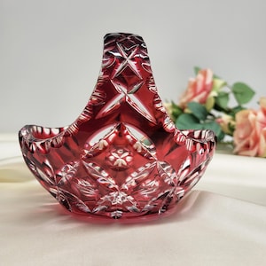 Könnte beinhalten: Ein rubinroter, geschliffener Glaskorb mit Henkel. Der Korb hat ein Diamanten- und Sternmuster. Der Griff ist gewölbt. Der Korb steht auf einer weißen Oberfläche. Im Hintergrund sind rosa Rosen zu sehen.