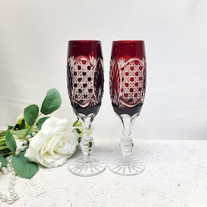 Könnte beinhalten: Zwei hohe Champagnergläser mit tiefroten Glaskelchen, die mit komplexen, klaren Schliffmustern in Rauten- und Kreuzschraffur versehen sind. Jedes Glas hat einen klaren Glasstiel mit einem runden Knoten und einen runden Fuß mit Sternschliff.