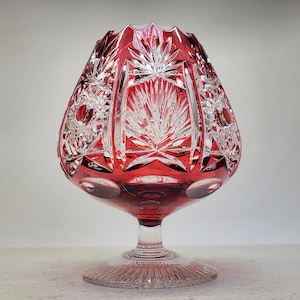 May include: A ruby red crystal brandy snifter with intricate cut glass patterns. The bowl-shaped glass sits atop a clear stem and a circular base. The cut glass design features starburst and fan-like patterns.