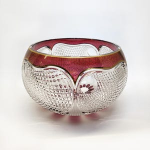 May include: A decorative glass bowl with a ruby red rim and gold trim. The bowl's body is clear, featuring a detailed, cut-glass pattern. The bowl is round and sits on a white surface.