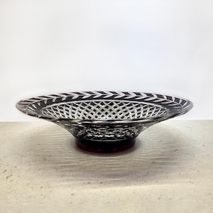 Può includere: Una ciotola di vetro rosso scuro con un motivo decorativo a foglie sul bordo e un disegno a griglia al centro. La ciotola poggia su una base corta e rotonda, su sfondo bianco.