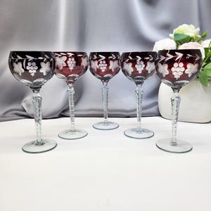 May include: Five ruby red wine glasses with clear, textured stems and bases. Each glass features a white etched grape and leaf design. The glasses are arranged in a row on a white surface, with a grey backdrop and a vase of flowers.