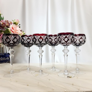 Può includere: Un set di sei calici da vino in cristallo con coppe rosso scuro caratterizzate da intricati motivi a diamante e a stella. Ogni calice presenta uno stelo trasparente sfaccettato con un dettaglio a rilievo e un'ampia base circolare.