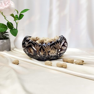 May include: A dark, deep purple cut crystal bowl featuring intricate starburst and geometric patterns, filled with unshelled peanuts. The bowl has a small circular sticker on its rim and sits on a smooth, draped fabric surface with scattered peanuts.