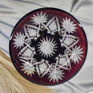 Könnte beinhalten: Ein rubinroter Servierteller aus Glas mit schwarzem Rand. Der Teller weist ein detailliertes, sternförmiges Muster aus klarem Glas auf. Das Design ist symmetrisch und aufwendig und erzeugt ein dekoratives und elegantes Aussehen. Der Teller wird wahrscheinlich zum Servieren von Speisen oder als Dekorationsstück verwendet.