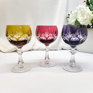 Op de afbeelding: Drie gekleurde kristallen wijnglazen. De glazen hebben een heldere steel en voet, met een decoratief geëtst ontwerp. De kommen zijn geel, rood en paars. De glazen staan op een wit oppervlak.