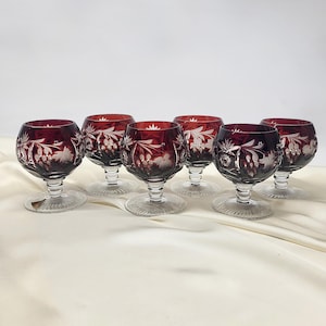 Può includere: Set di sei calici da vino in cristallo rosso rubino. Ogni calice presenta una coppa rotonda con disegni floreali incisi e un gambo e una base chiari e dettagliati. I calici sono disposti su una superficie bianca.