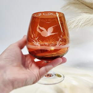 Può includere: Bicchiere da brandy color ambra con un disegno inciso di un uccello in volo sull'acqua e canne. Il bordo ha un motivo a spirale decorativo. Lo stelo è in vetro trasparente.