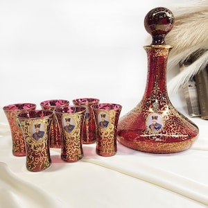 Può includere: Un set di caraffa in vetro rosso rubino con sei bicchierini coordinati. Il set presenta filigrane dorate e ritratti. La caraffa ha una base rotonda e un collo lungo con un tappo rotondo. I bicchieri sono alti circa 7,6 cm.