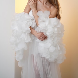 May include: A woman wearing a white, strapless gown with large, ruffled sleeves. The sleeves are made of layered, petal-like fabric. The gown has a sheer, flowing skirt. The background is a gradient of white to yellow.