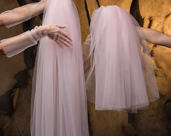 May include: Two sheer, pale pink veils are held up by unseen hands. The veils are long and flowing, with a soft, delicate texture. The background is a textured, neutral-toned wall.