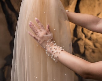 May include: A cream-coloured bridal veil made of sheer fabric, decorated with scattered pearls. The veil is held up, displaying its delicate texture and the subtle sparkle of the pearls. Sheer gloves with pearl accents are also visible.