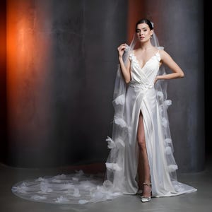 May include: A white satin wedding dress with a high slit and a long train. The dress features a V-neckline and floral embellishments. A sheer veil with floral accents drapes from the shoulders. The model is wearing silver heels.