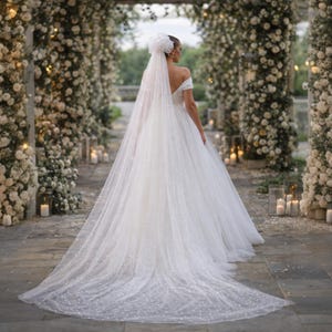 May include: A white bridal gown with a long train and a veil. The dress features an off-the-shoulder neckline and a full skirt. The veil is adorned with small details. The setting is outdoors, with floral arches and candles.