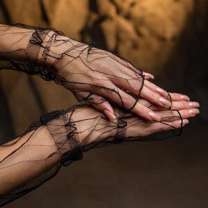 May include: Black, fingerless gloves crafted from fine, sheer lace. The gloves feature a ruffled trim and a delicate, web-like pattern. The hands are posed to showcase the gloves, with long, manicured nails. The backdrop is a warm, neutral shade.